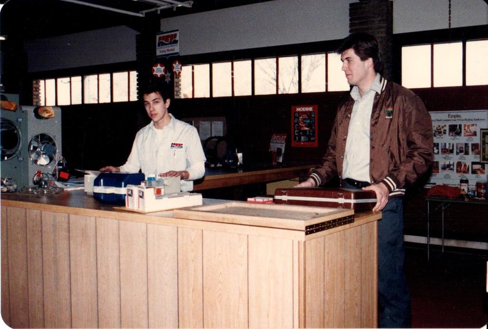 Two young men at a wood paneled parts counter in the 1980s with heating equipment displays behind them