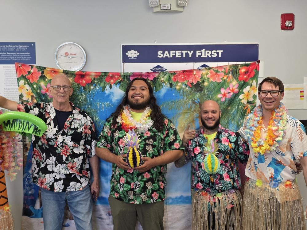 Four employees in Hawaiian shirts and leis in front of a tropical backdrop and a Safety First banner
