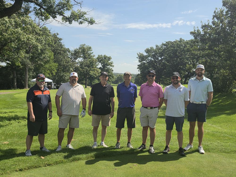 Seven golfers lined up on a tee box on a clear summer day