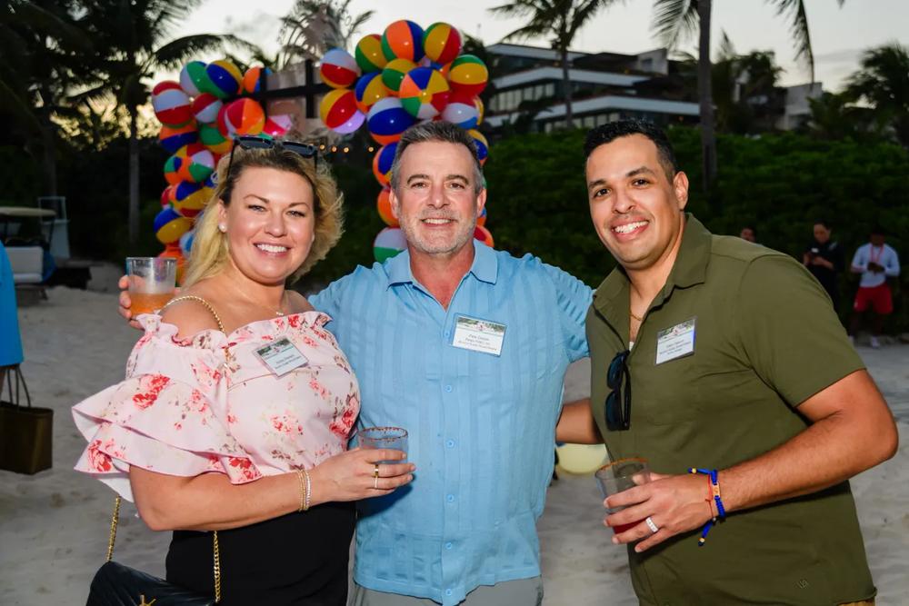 Three attendees with drinks in front of a beach ball arch at the dealer trip