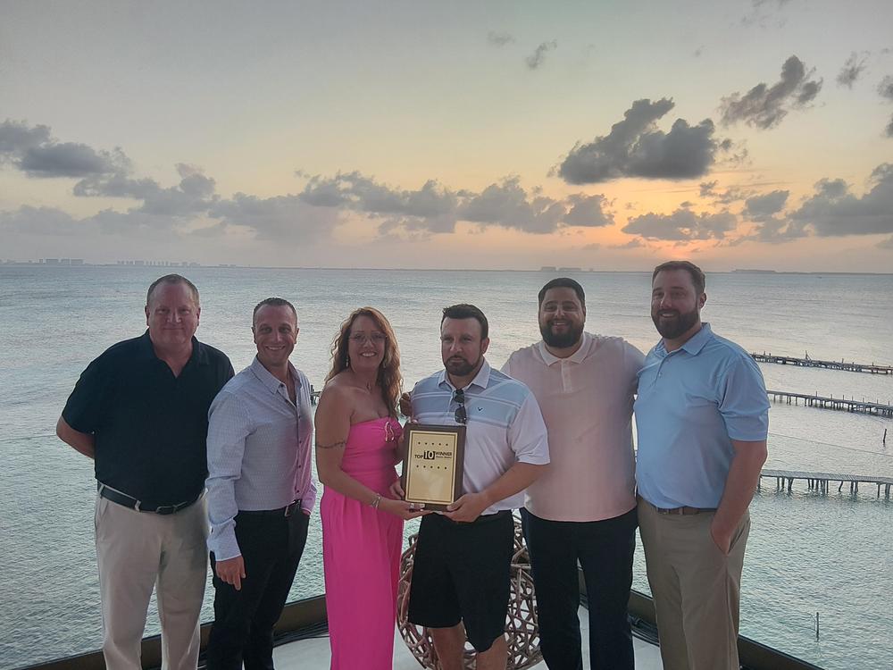 Six people holding a Top 10 Winner plaque on a terrace overlooking the water at sunset